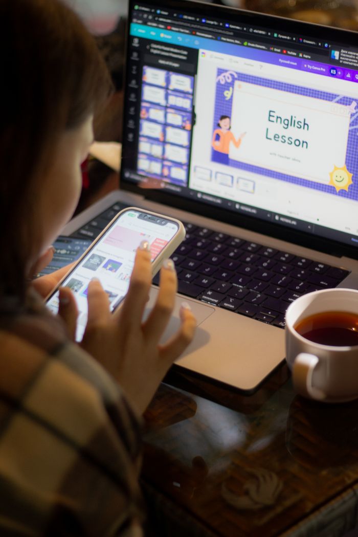 Adult woman multitasking, using laptop and smartphone while studying English with tea on a table.